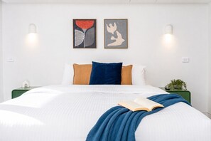 A neatly made bed with a blue blanket and an open book, two framed artworks on the wall, and bedside lamps. - The Pavilion | Records, Space + MCG Moments Away! (Richmond)