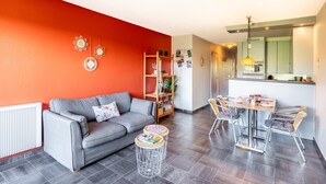 Un salon moderne avec un canapé gris, une table à manger avec des chaises, un coin cuisine et un mur d’accent rouge.