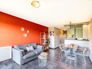 A modern living room with a grey sofa, a dining table with chairs, a kitchen area, and a red accent wall. - Charming apartment close to the the beach (Sint-Idesbald)