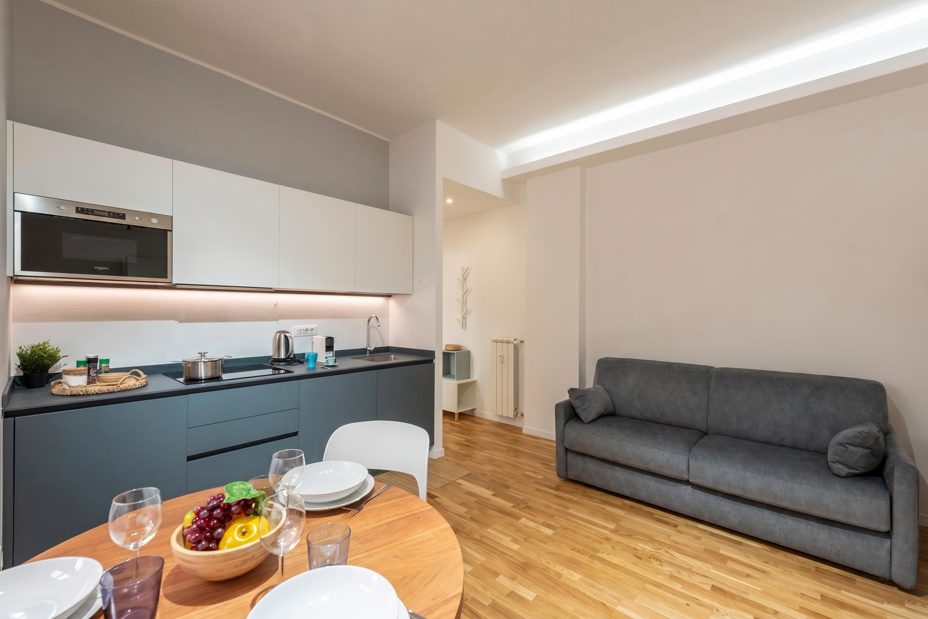 Eine moderne Küche mit Einbauofen, ein Essbereich mit Holztisch und Stühlen sowie ein graues Sofa im Wohnbereich.