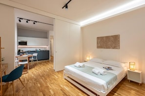 A modern hotel room with a bed, bedside lamps, a desk, and a view into the kitchen area.