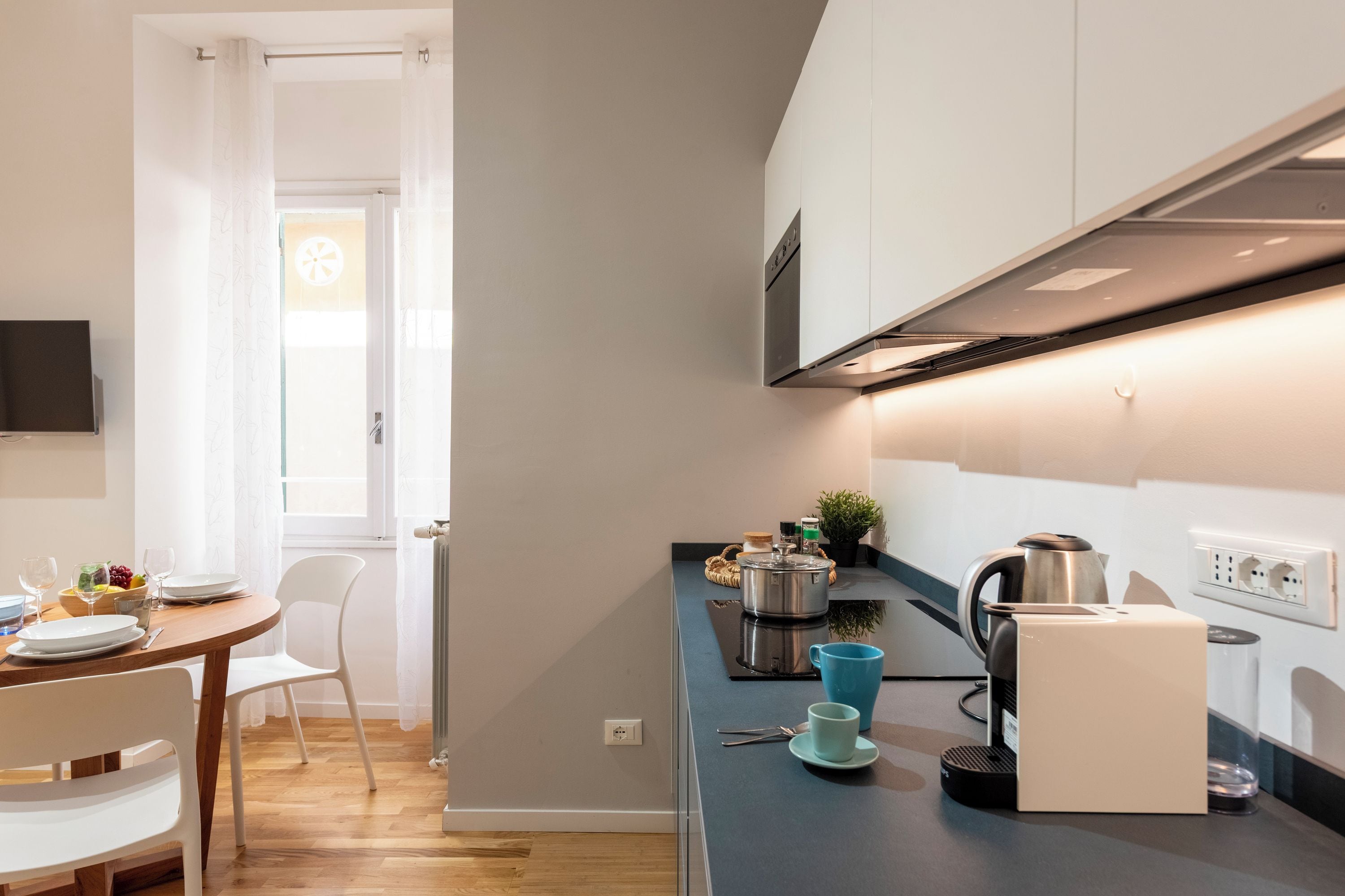 A modern kitchen with a dining area, a kettle, a coffee machine, and a window with curtains.