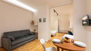 A modern living room with a gray sofa, a wooden dining table with chairs, a TV mounted on the wall, and a small potted plant. - Maison Columbus in the Heart of Genova (Genoa)