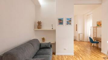 Un salon moderne avec un canapé gris, un plancher en bois, un mur blanc orné d’œuvres d’art et un bureau avec une chaise.