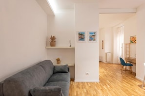 A modern living room with a grey sofa, wooden flooring, a white wall with artwork, and a desk with a chair.