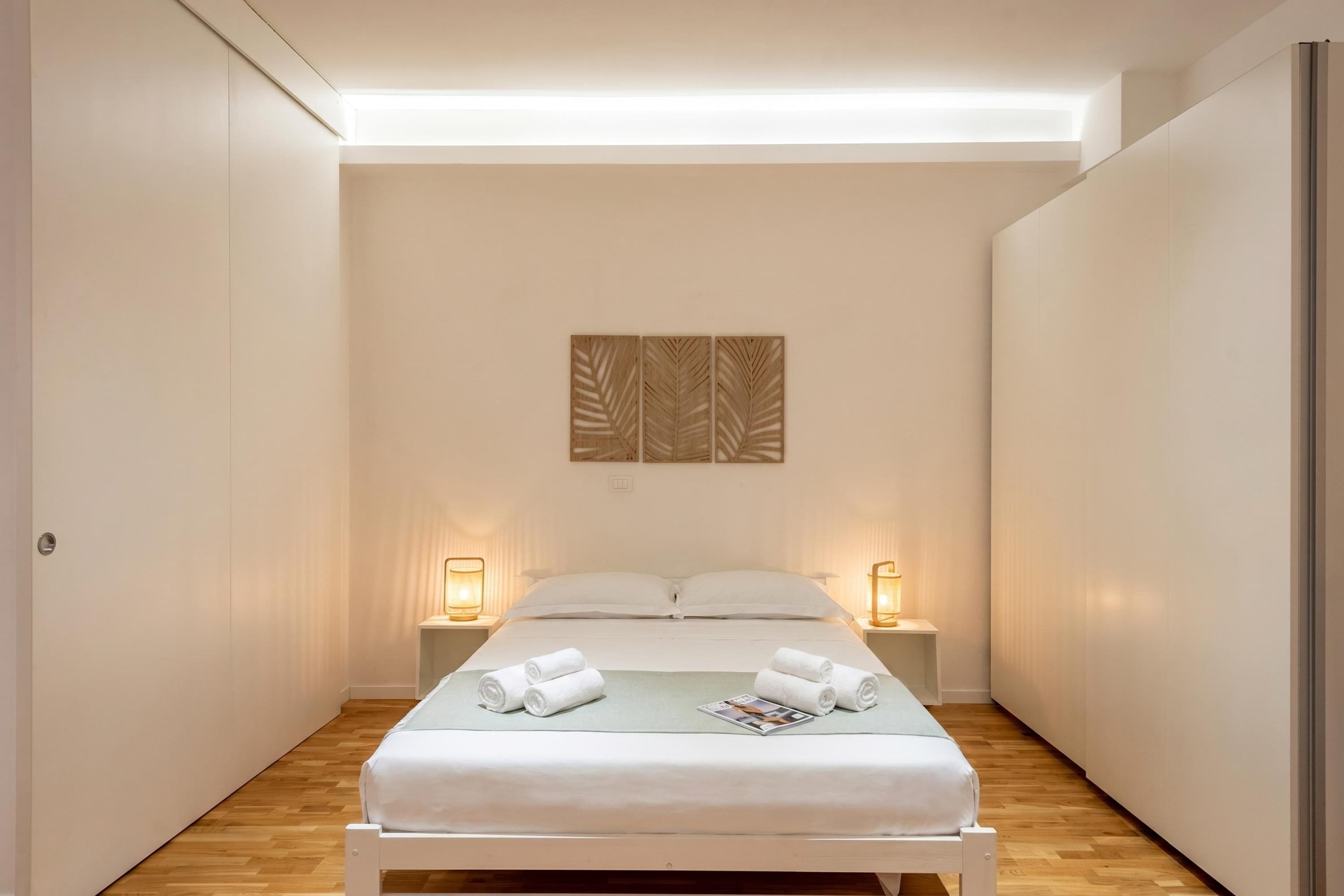 Ein schmales Hotelzimmer mit einem Bett, Nachttischlampen und einem an der Wand hängenden Kunstwerk.