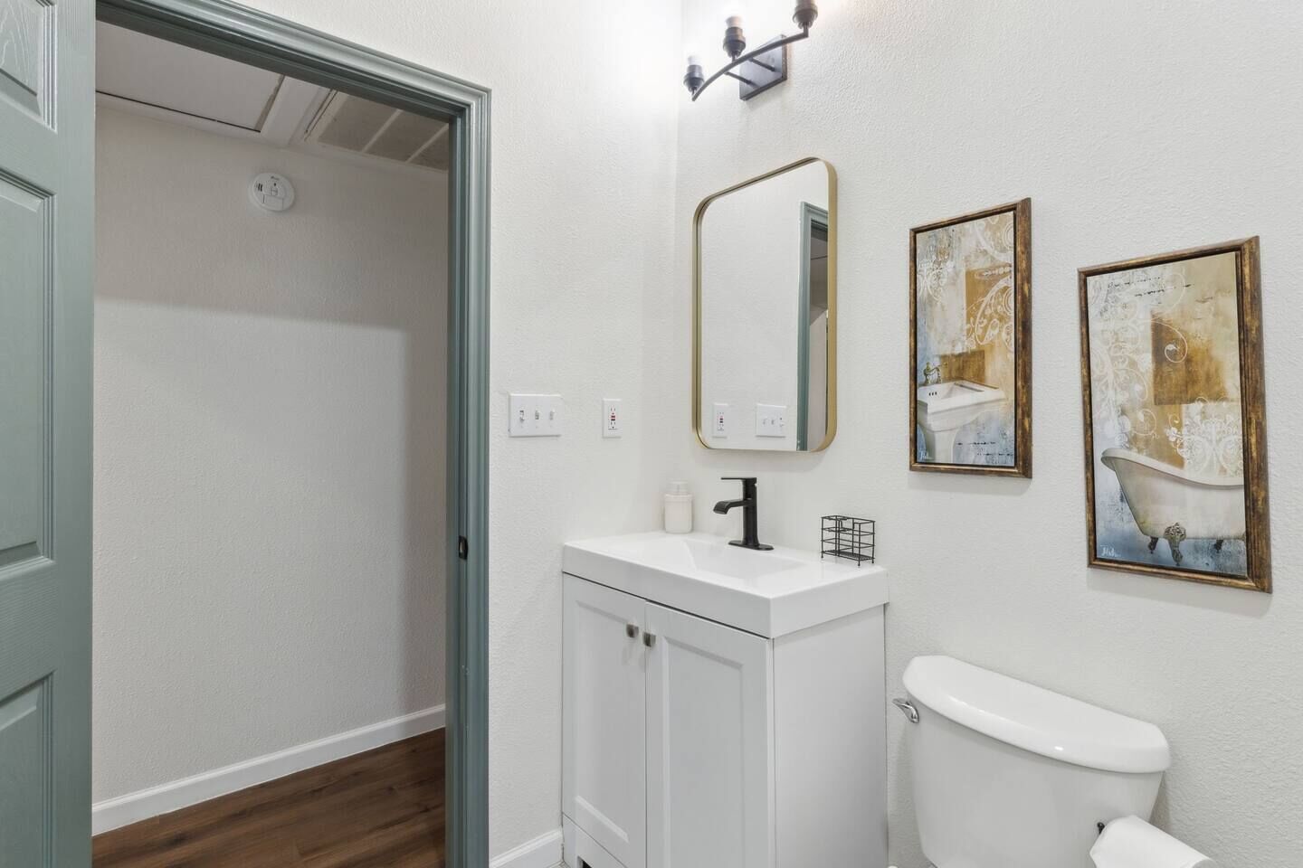 A bathroom with a toilet, a sink, and two mirrors. There are two framed artworks on the wall.