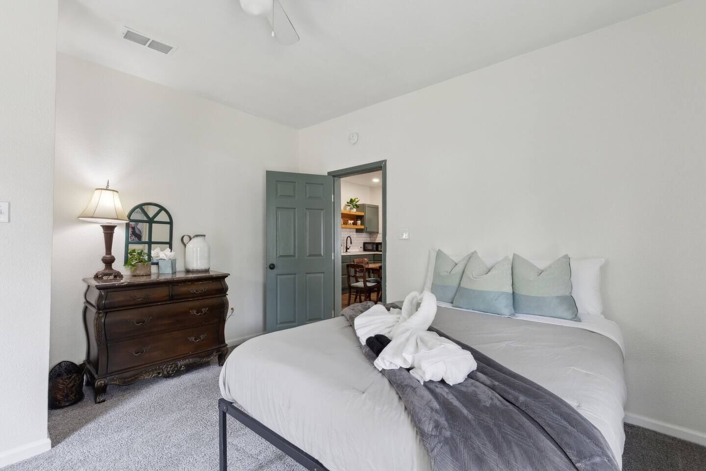 A bedroom with a bed, dresser, lamp, and a door leading to a kitchen area.