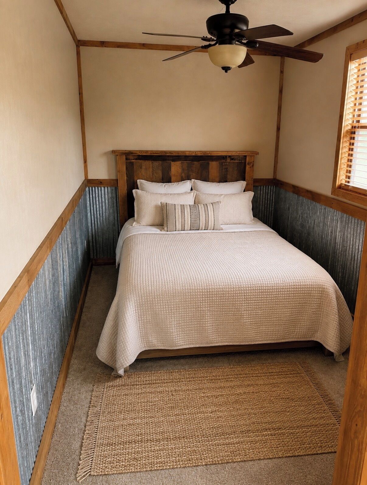 A bedroom with a wooden headboard, a bed with a beige quilt, a woven rug, and a ceiling fan.