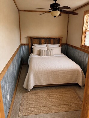 A bedroom with a wooden headboard, a bed with a beige quilt, a woven rug, and a ceiling fan.