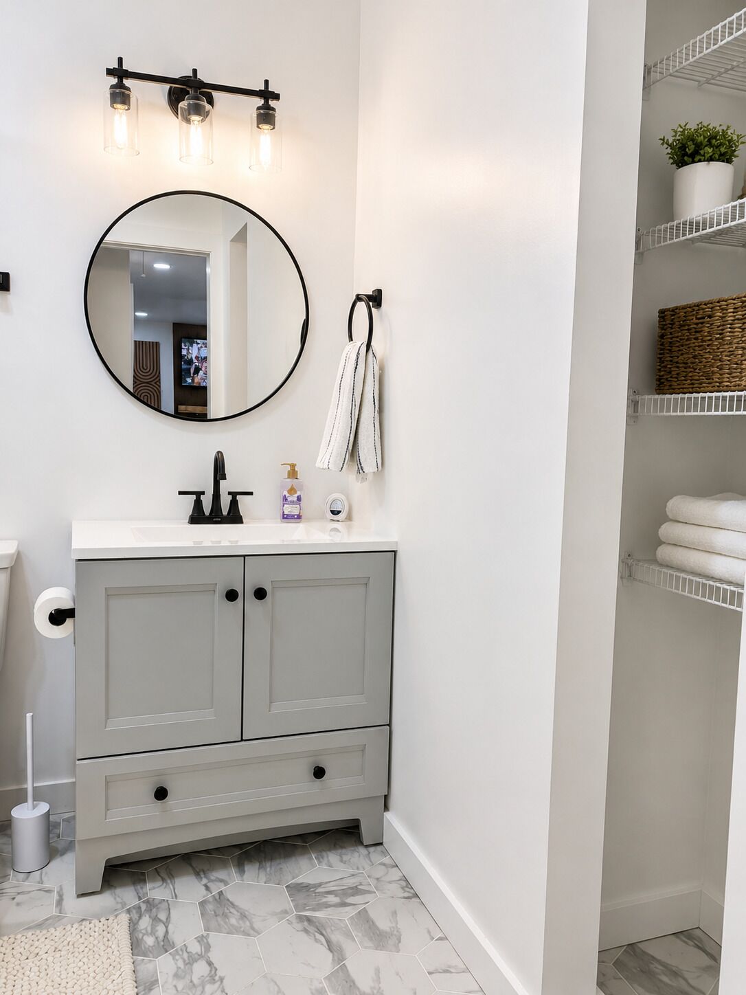Un baño con un tocador gris, un espejo redondo, un perchero, un dispensador de papel higiénico y una repisa con una planta y toallas.