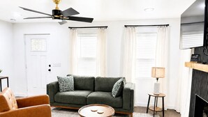 Uma sala de estar com sofá, poltrona, mesa de centro redonda, ventilador de teto e lareira.