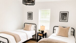 Um quarto com duas camas de solteiro, uma mesa de cabeceira com abajur, janela com persianas e quadros nas paredes.
