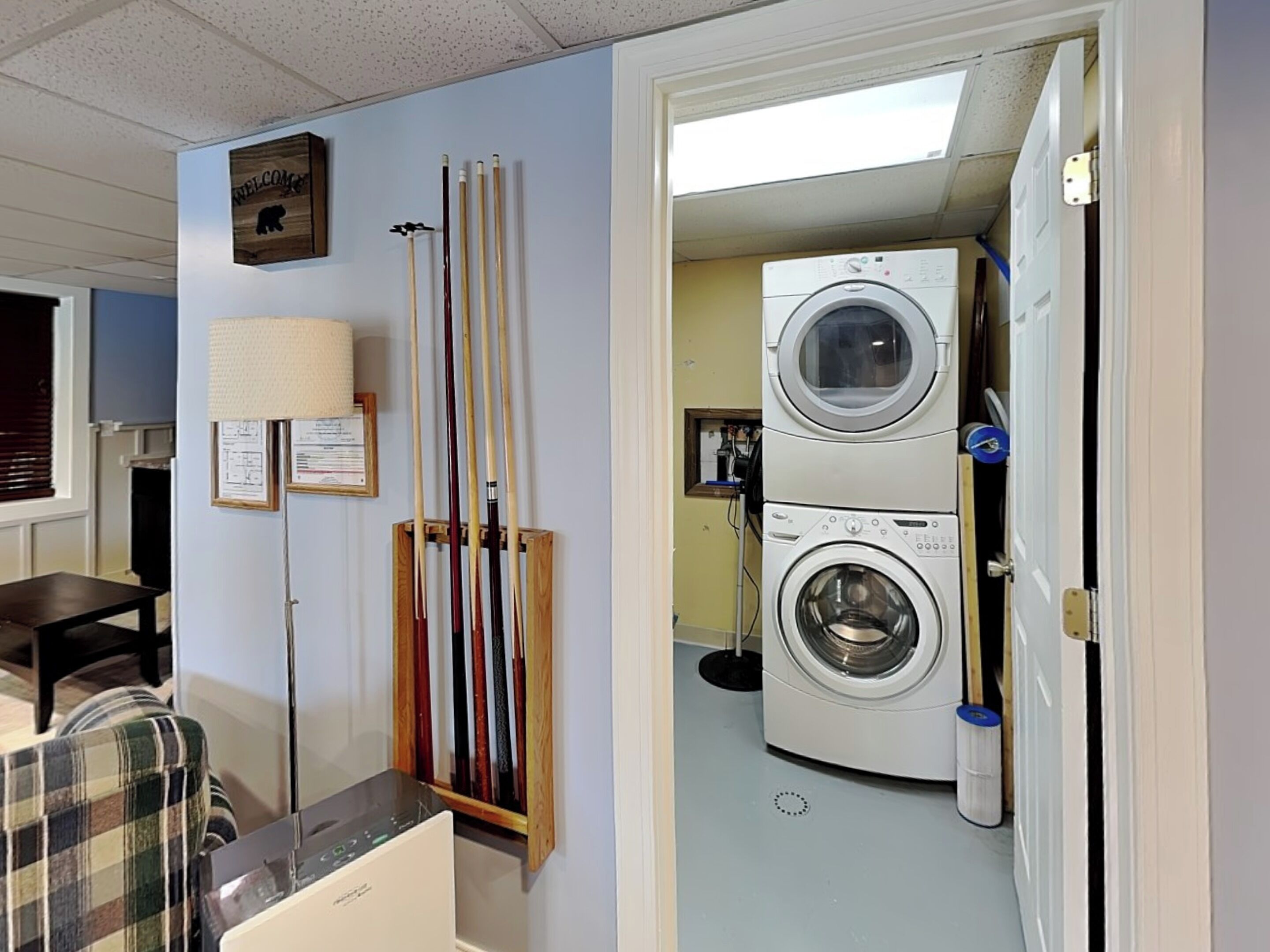 A room with a pool table, cue sticks, a washing machine, and a plaid chair.