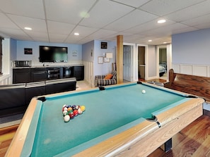 Una mesa de billar con bolas, un televisor, una zona de cocina y un área de descanso con sofá y sillones.