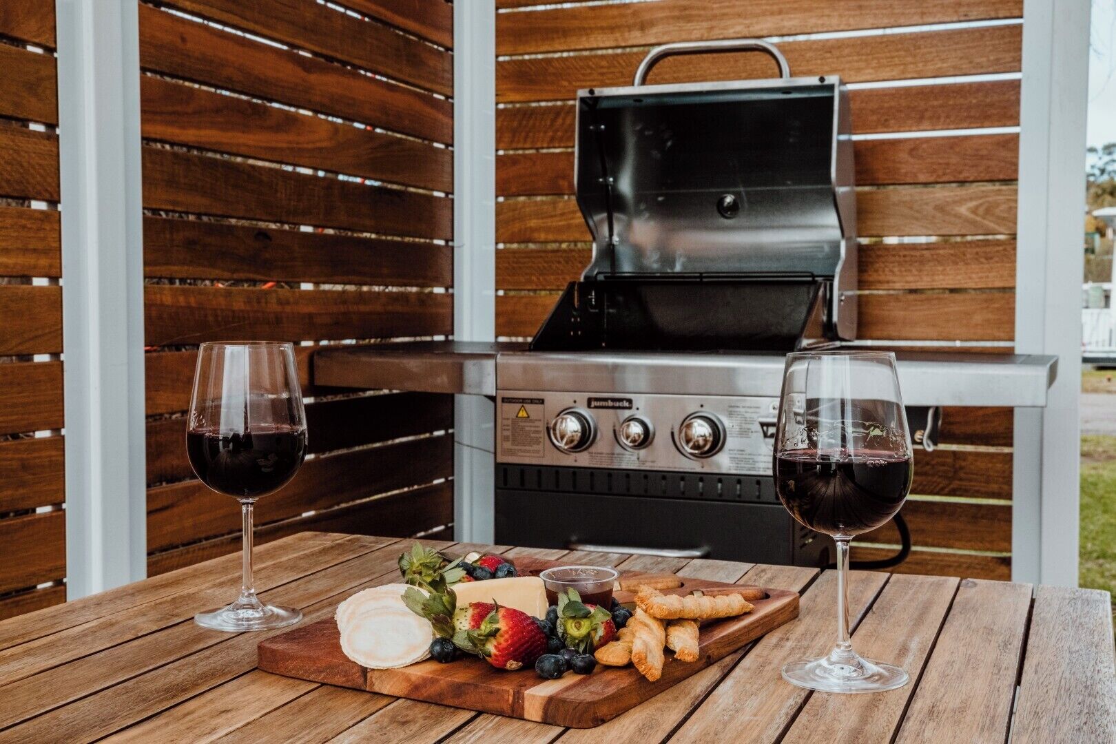An outdoor grill with a wooden table in front, two glasses of red wine, and a variety of food including cheese, strawberries, and pastries.