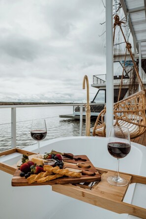 A table with a glass of red wine, a wooden board with fruits and pastries, and a view of the water.