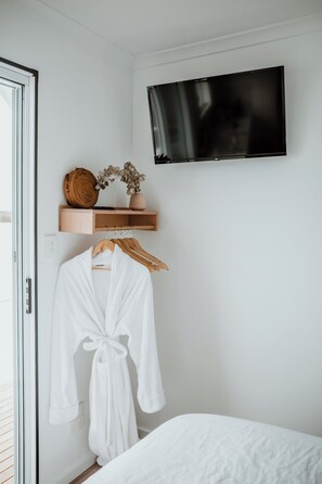 A bedroom with a bed, a TV mounted on the wall, a wooden shelf with decorative items, and a white robe hanging on a wooden hanger.