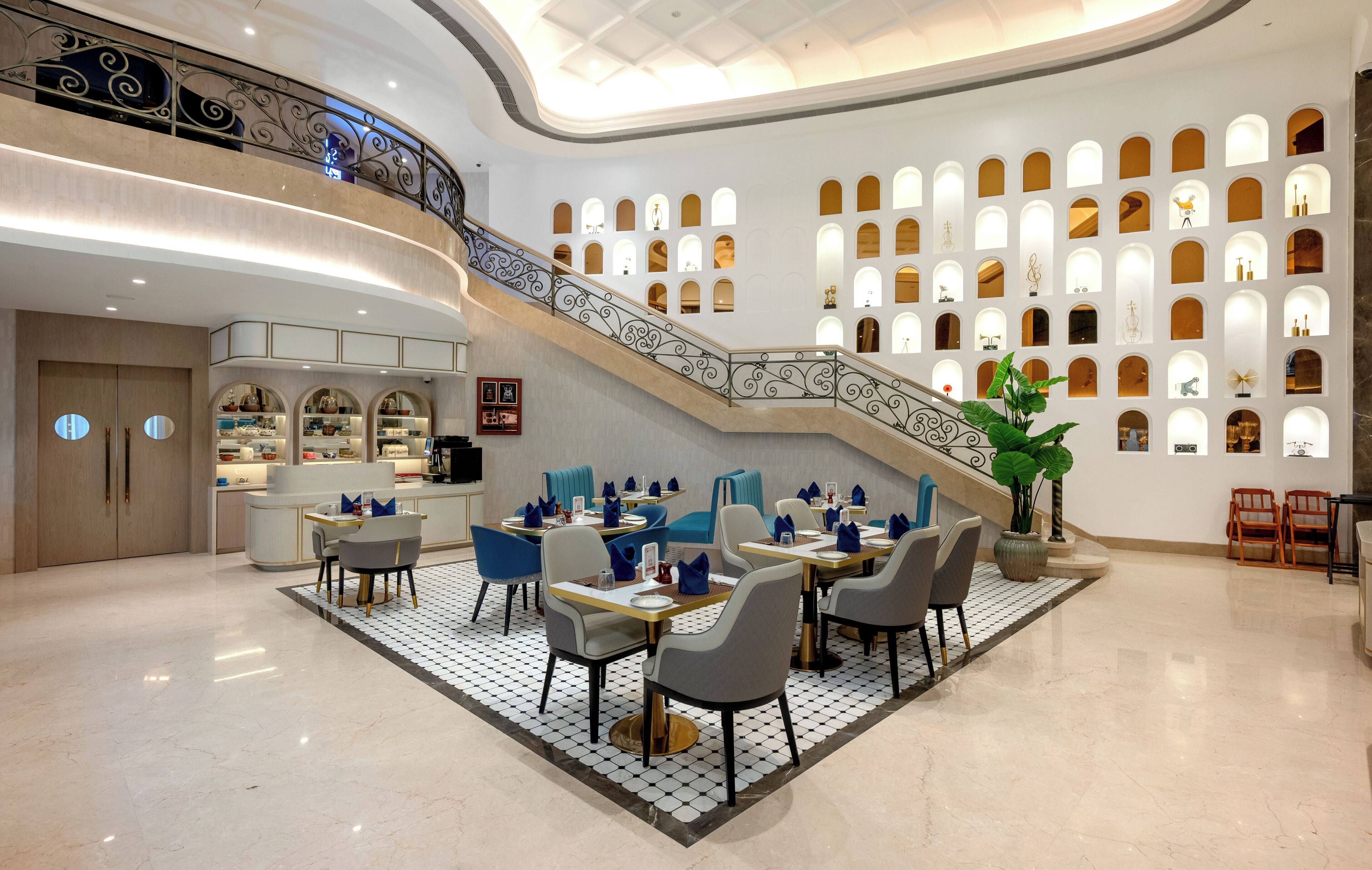 Un vestíbulo espacioso con una imponente escalera, un comedor con sillas y mesas modernas, y una pared con numerosas ventanas pequeñas.