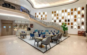 A spacious lobby with a grand staircase, a dining area with modern chairs and tables, and a wall with numerous small windows. - Hotel Express Inn Heritage (Chhatrapati Sambhajinagar)