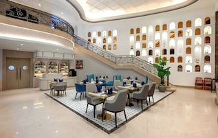 Un vestíbulo espacioso con una imponente escalera, un comedor con sillas y mesas modernas, y una pared con numerosas ventanas pequeñas.