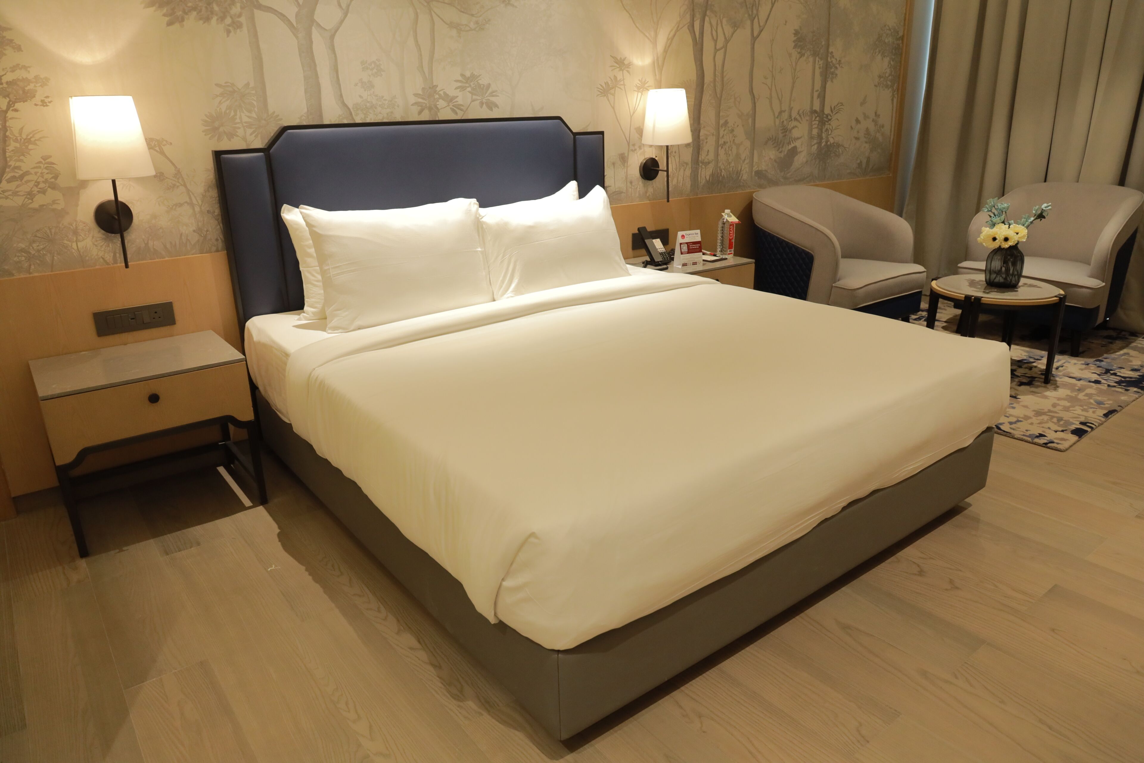 A hotel room with a large bed, bedside tables, a chair, a small table with a vase, and wall-mounted lamps.