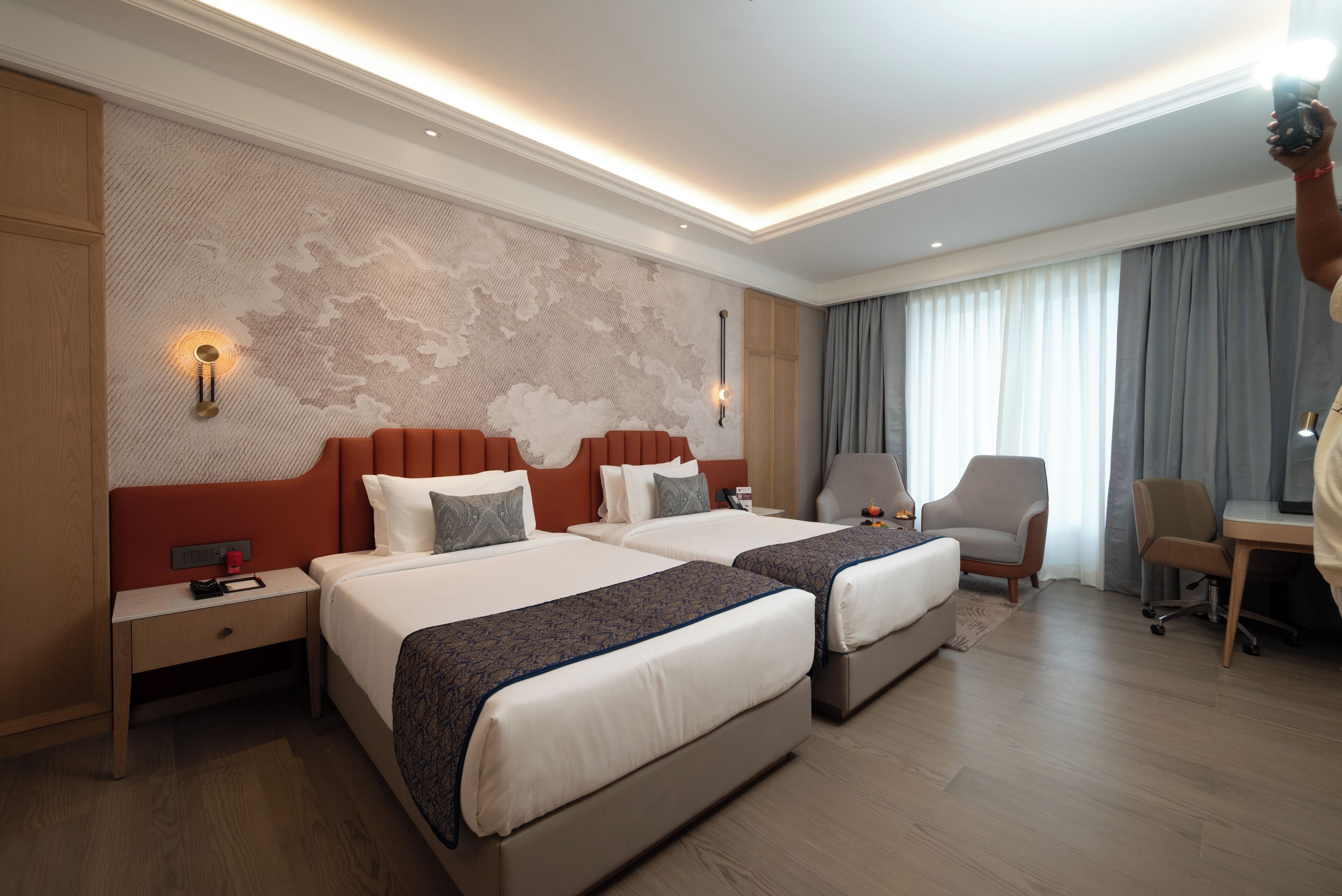 Habitación de hotel con dos camas, un escritorio, una silla y un ventanal con cortinas.