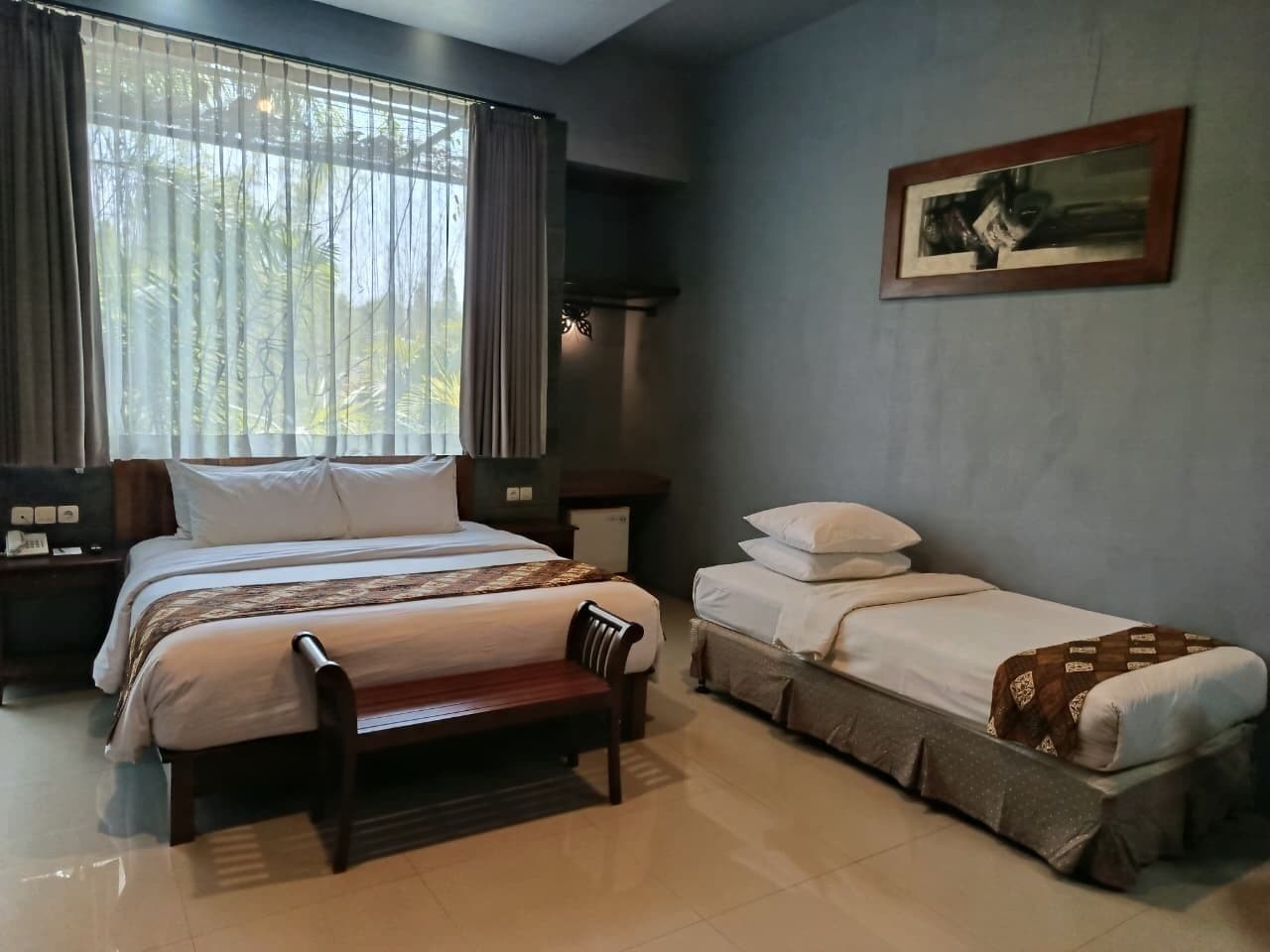 A bedroom with a large bed, a smaller bed, a bench, a nightstand, and a wall-mounted picture.