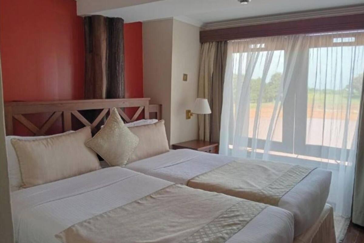 Habitación de hotel con dos camas, cabecero de madera, mesita de noche, lámpara y vistas al exterior a través de cortinas traslúcidas.