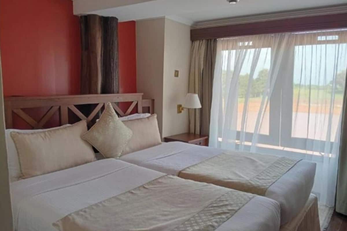 A hotel room with two beds, a wooden headboard, a nightstand, a lamp, and a view of the outdoors through sheer curtains.