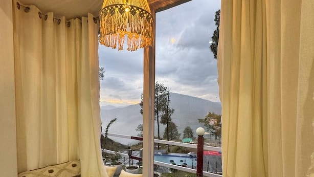 A room with a bed against a window, curtains, a hanging light fixture, and a view of a mountainous landscape.