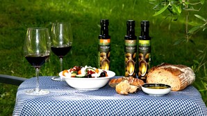 Una mesa con mantel a cuadros, un bol de ensalada, dos copas de vino tinto, tres botellas de aceite de oliva, un pan y un recipiente pequeño con salsa.