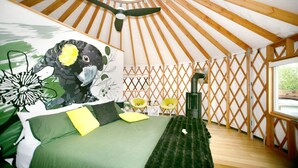 Una yurtas con una cama, una manta verde, un cojín amarillo, techo de madera y una ventana.