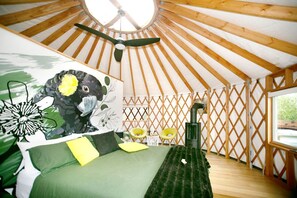 Une yourte avec un lit, une couverture verte, un coussin jaune, un plafond en bois et une fenêtre.
