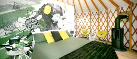 A tent-style room with a bed, a wall mural, a small table with a chair, and a wood-burning stove.