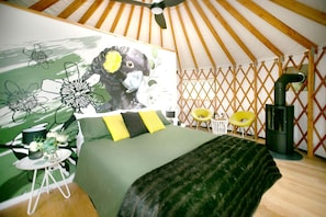 A tent-style room with a bed, a wall mural, a small table with a chair, and a wood-burning stove.