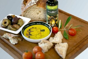 A wooden board with a bowl of yellow dip, olives, cherry tomatoes, bread, and a bottle of olive oil.