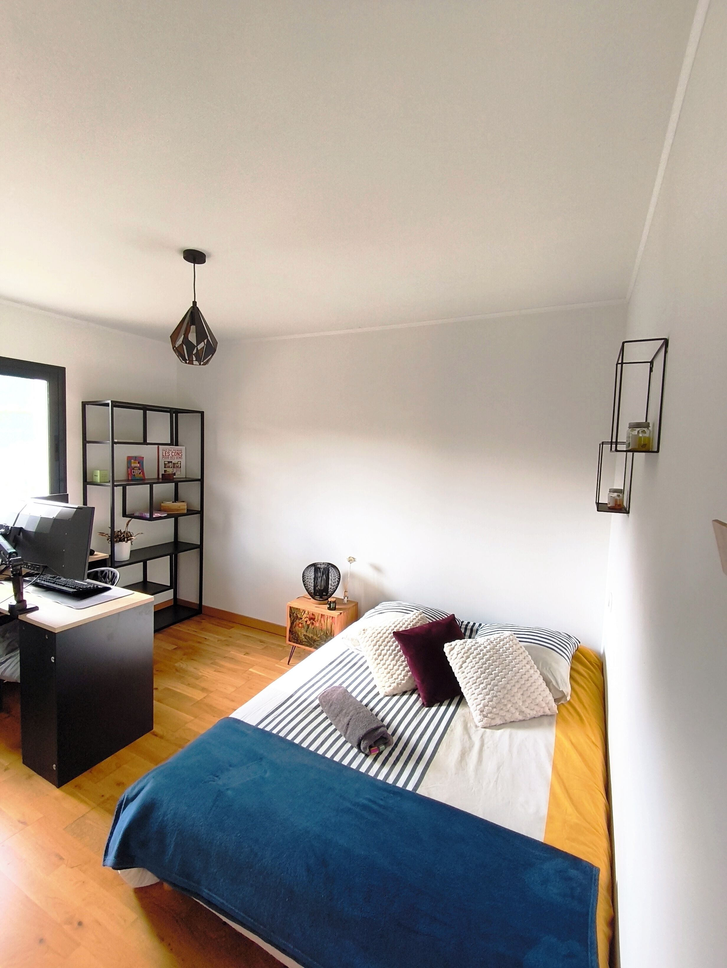 A bedroom with a bed, a desk with a computer, a bookshelf, and a hanging light fixture.