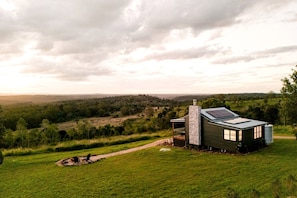 Ein modernes Haus mit einem großen Panoramafenster, einem Steinkamin und einer Solaranlage auf dem Dach, umgeben von einer üppigen grünen Landschaft.