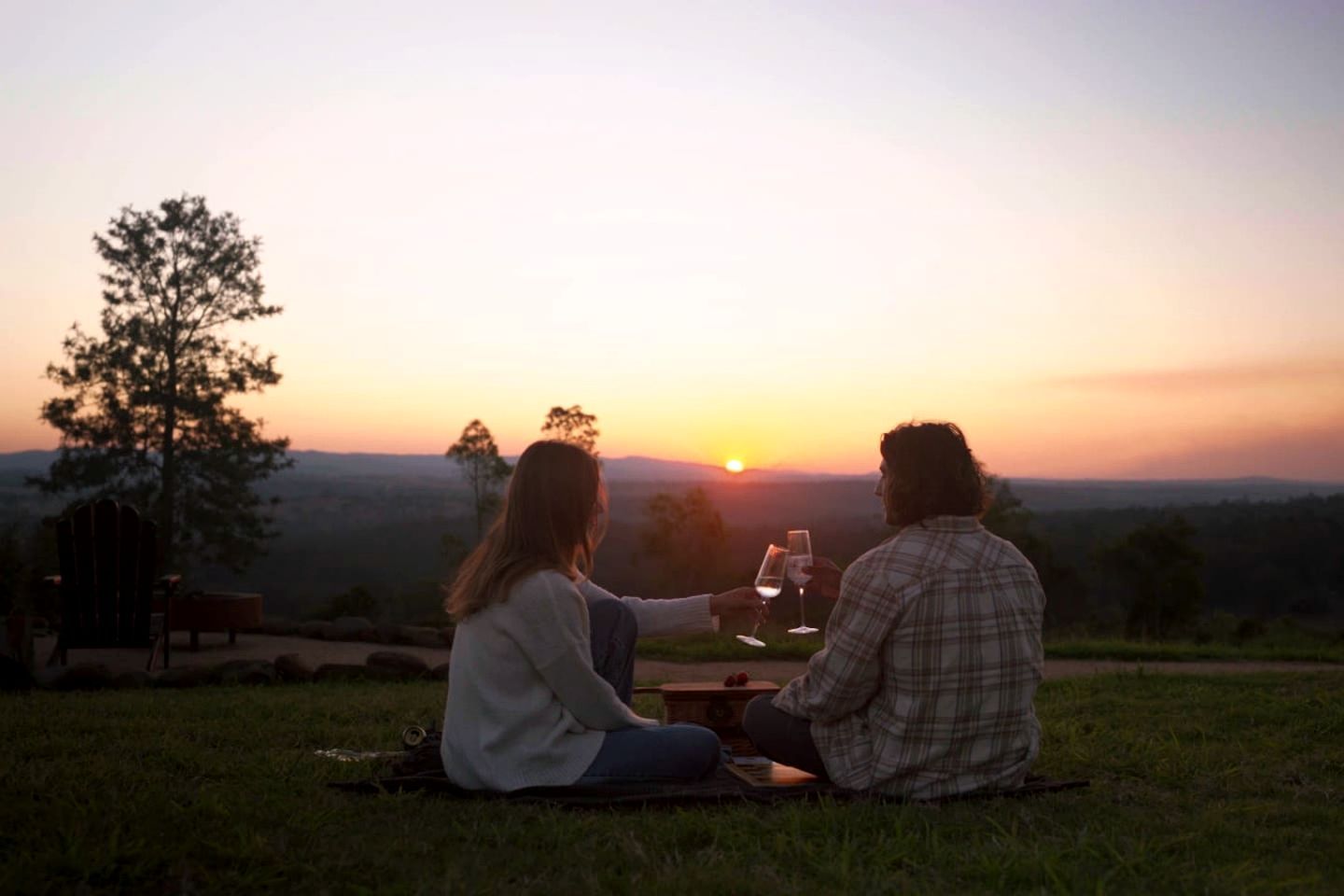Two people sitting on a blanket outdoors during sunset, with a view of the landscape.