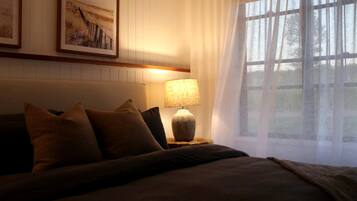 Un dormitorio con una cama, almohadas, una lámpara, un ventanal con cortinas traslúcidas y obras de arte enmarcadas en la pared.