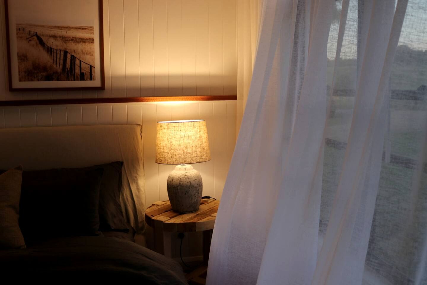 Ein Schlafzimmer mit Bett, Nachttischlampe, einem gerahmten Bild an der Wand und einem Fenster mit Vorhängen.