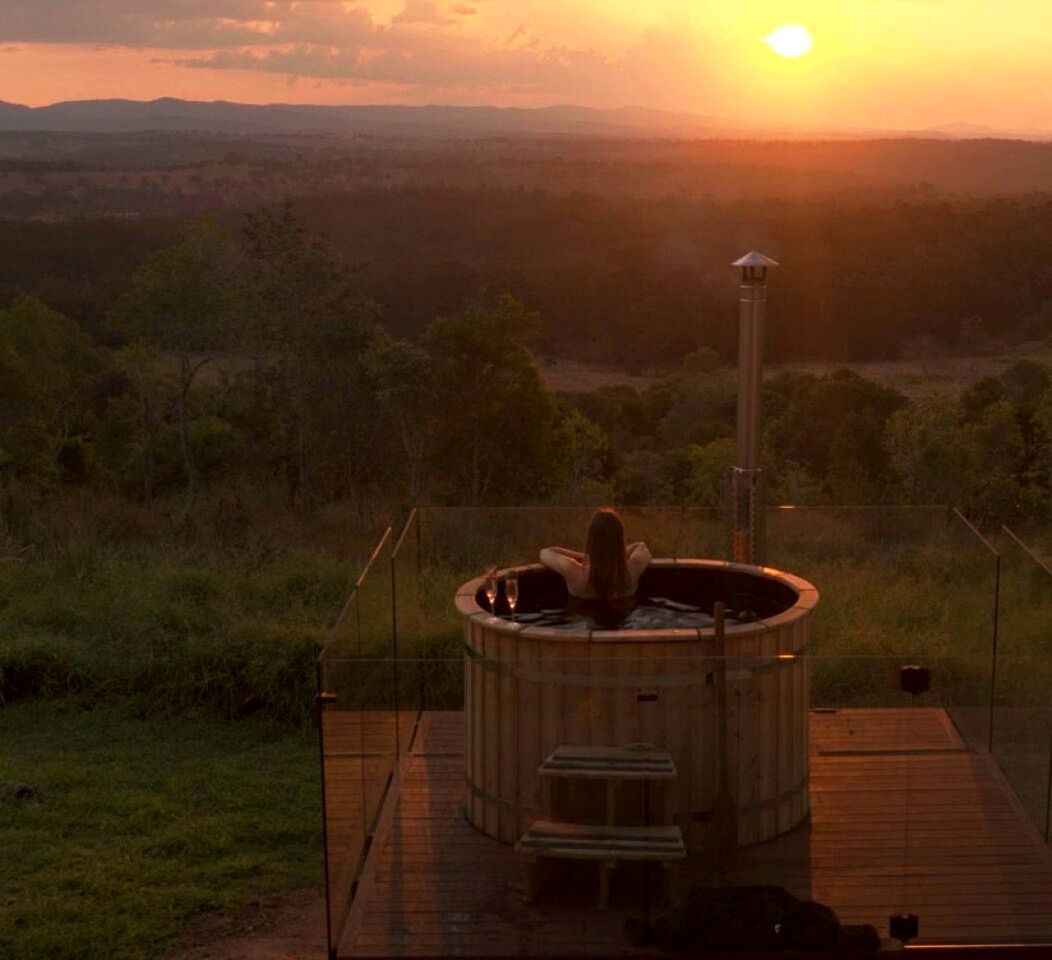 A hot tub with a wooden deck and a view of a sunset over a hilly landscape.