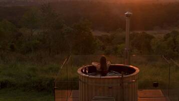 A hot tub with a wooden deck and a view of a sunset over a hilly landscape.