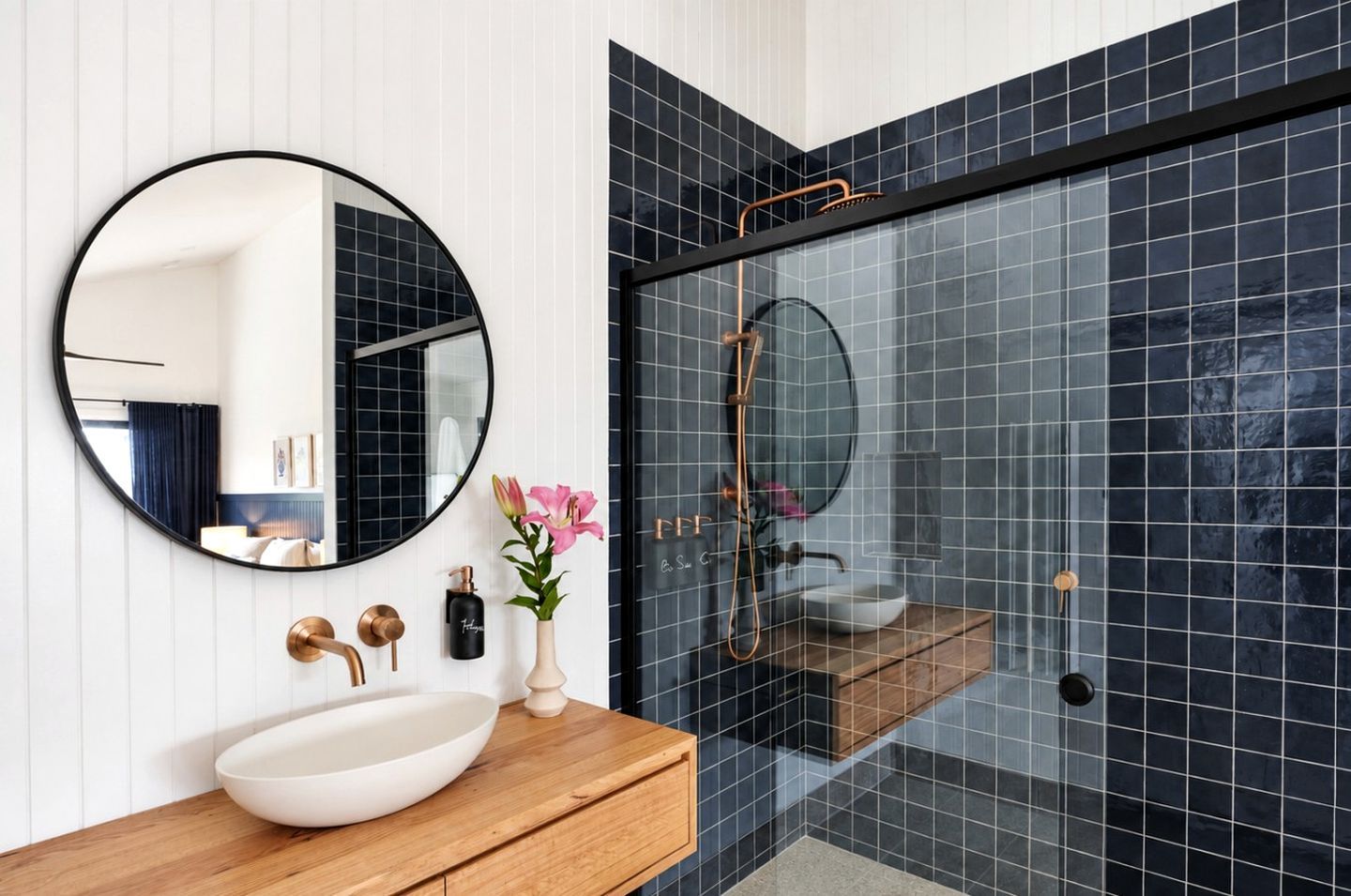 A modern bathroom with a round mirror, a wooden vanity, a white sink, and a glass shower enclosure.