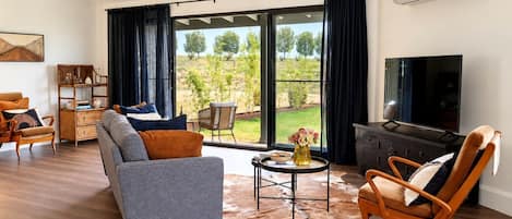 Ein modernes Wohnzimmer mit Sofa, Sesseln und Couchtisch. Es gibt einen Fernseher und ein großes Fenster mit Vorhängen.