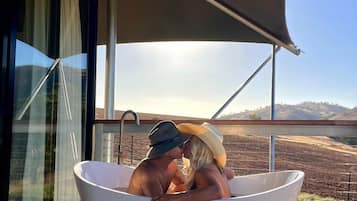 Eine Person entspannt in einer großen weißen Badewanne auf einer Holzdielen-Terrasse mit Blick auf einen Weinberg und Hügel.