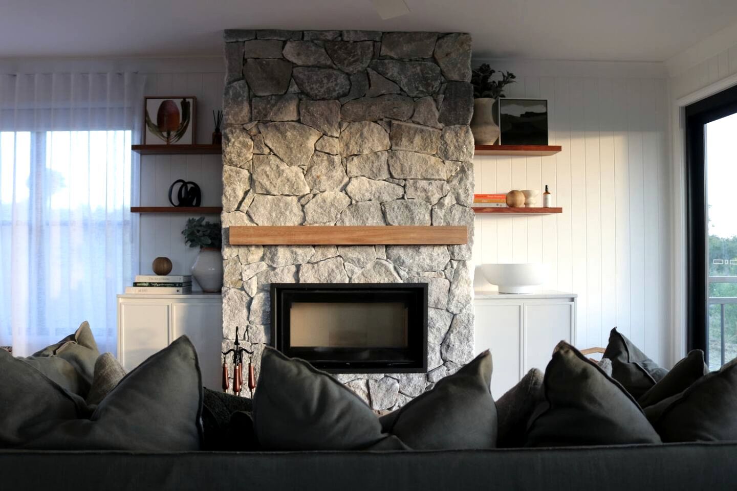 A living room with a stone fireplace, a flat-screen TV, a sofa with cushions, and a large window.