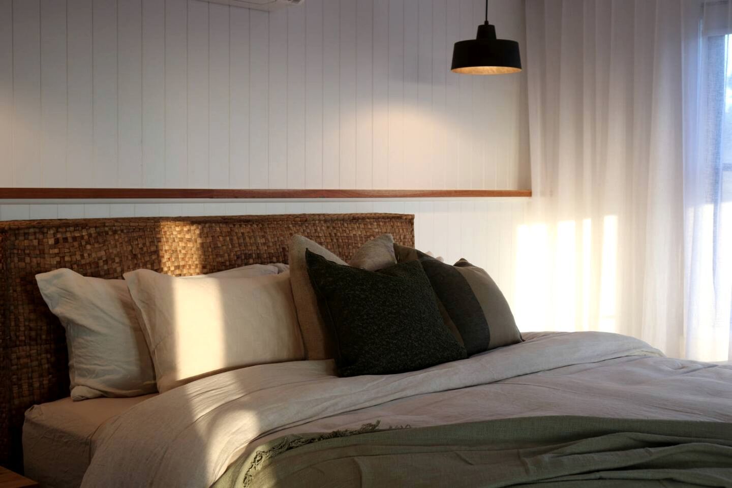 Una cama bien hecha con un cabecero tejido, una lámpara colgante y cortinas traslúcidas.
