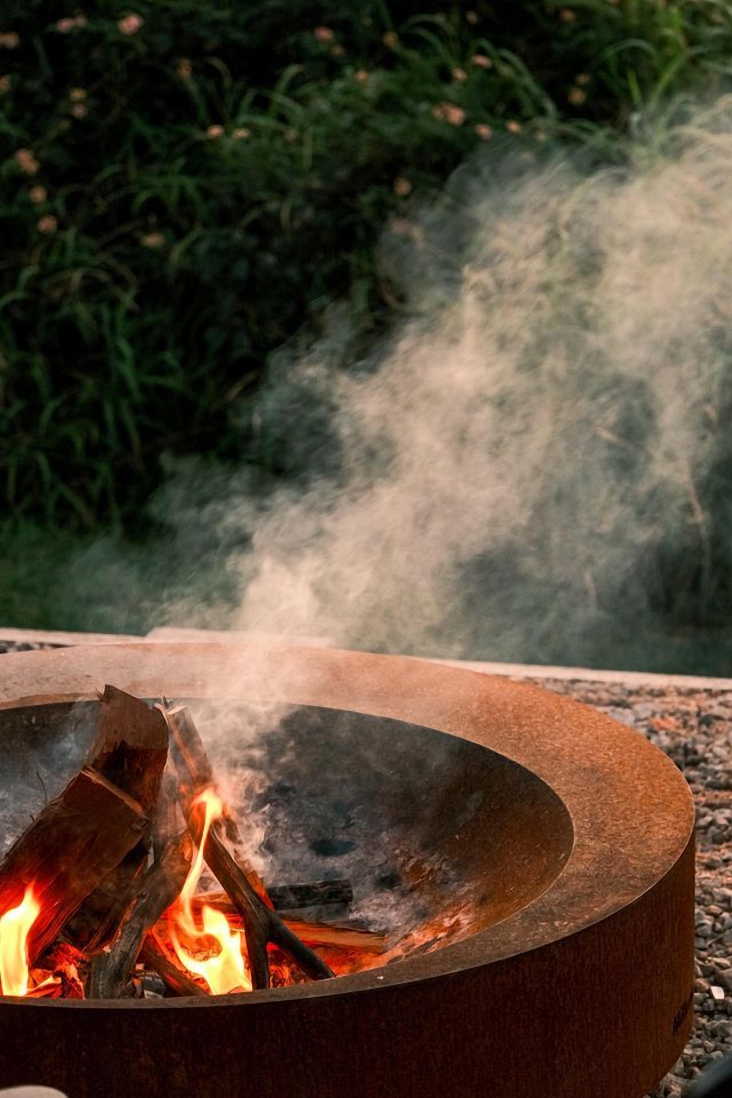A circular metal fire pit with burning logs and smoke.
